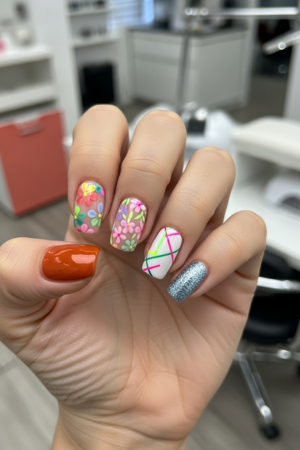 “Close-up of a straight flat hand with seasonal nail designs, including burnt orange autumn nails, pastel floral spring nails, neon summer accents, and glittery silver-blue winter nails. Bright lighting with a softly blurred modern nail salon background.”